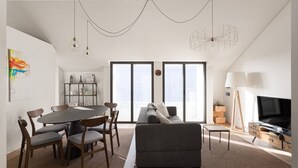 A modern living room with a grey sofa, dining table with chairs, a TV, and a large window with blinds. - JOIVY Spacious flat in Bairro Alto (Lisboa)