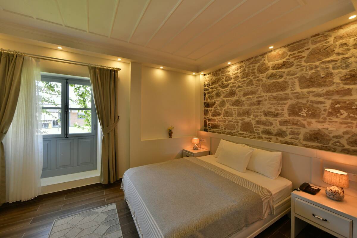 Ein Schlafzimmer mit Steinwand, einem Bett mit weißen Leinen, einem Nachttisch mit Lampe und einem Fenster mit Vorhängen.