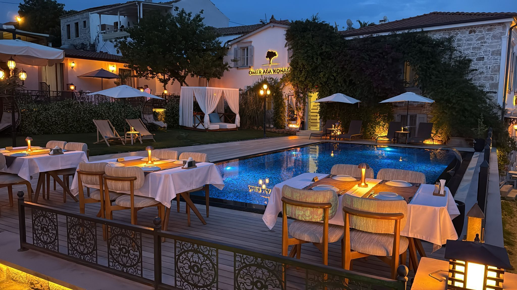 Un área de comedor al aire libre con mesas y sillas alrededor de una piscina, un letrero que dice "OMER AGA KONAG" y un edificio con fachada de piedra.