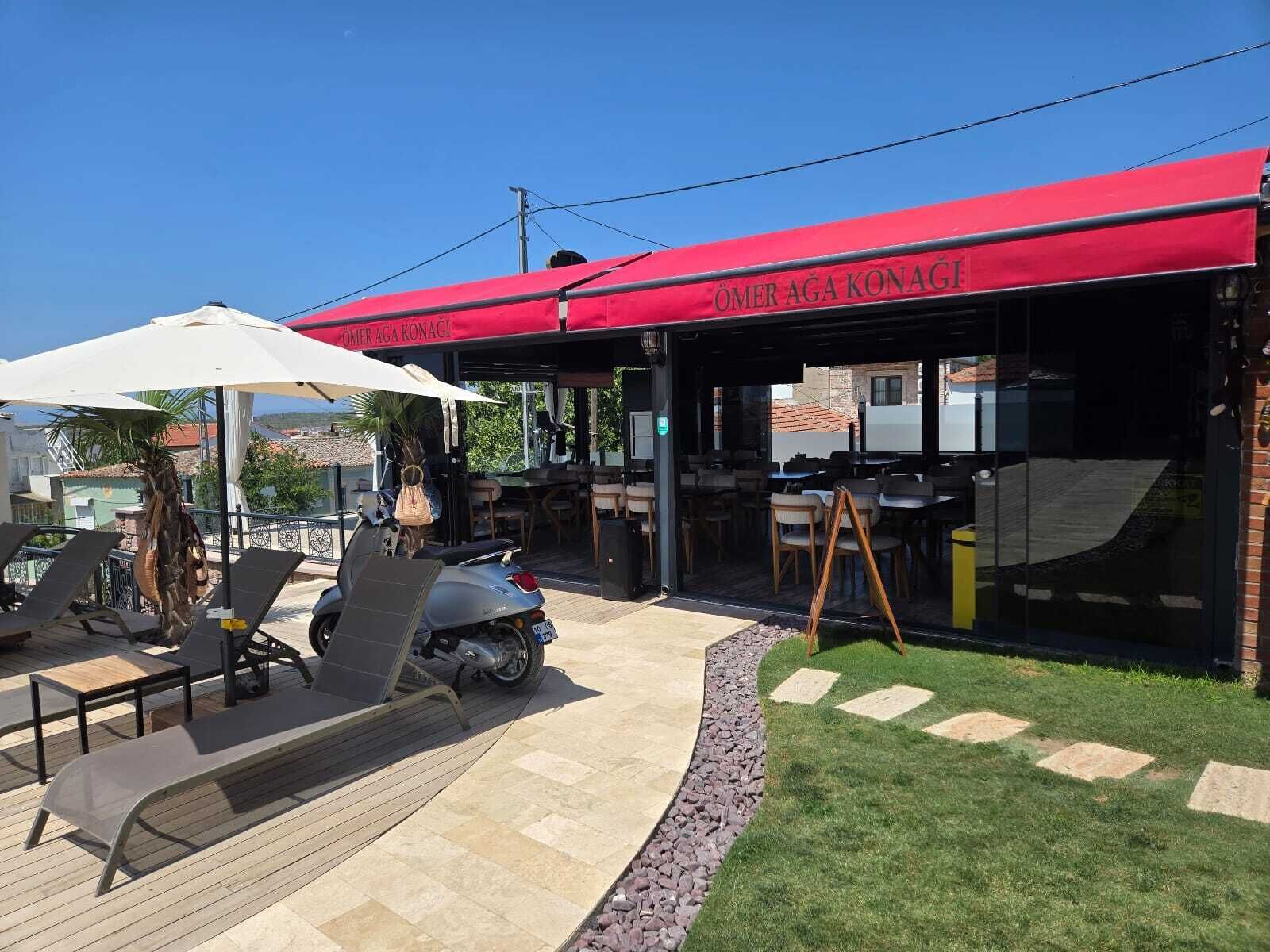 Área de estar ao ar livre com guarda-sóis, uma scooter e um restaurante com toldo vermelho.