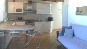 Una cocina moderna con armarios blancos, encimera de mármol y horno empotrado. Hay una zona de comedor con mesa y sillas, y un sofá en la sala de estar.
