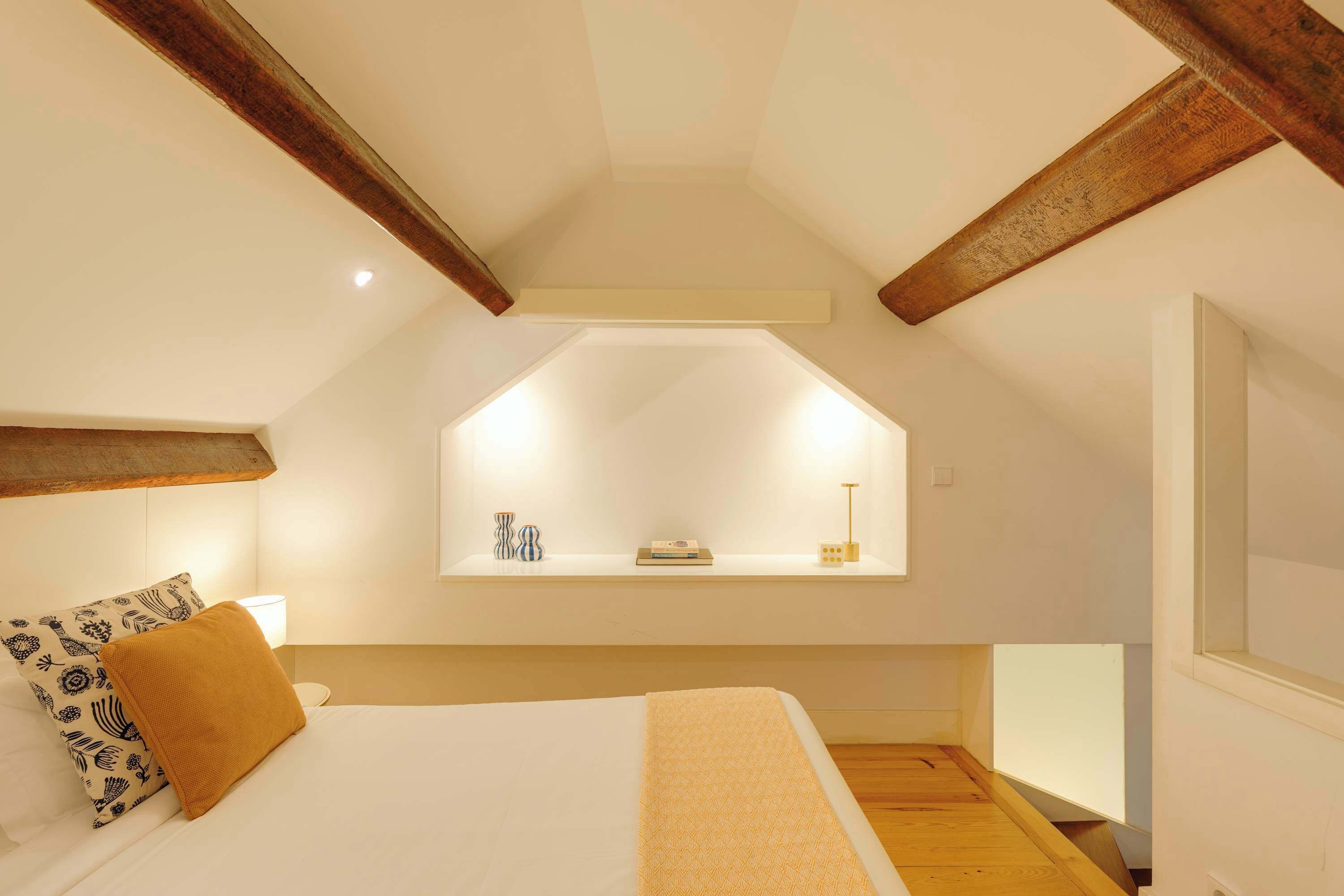 A bedroom with a sloped ceiling, a bed with white linens, a brown throw blanket, a bedside table with a lamp, and a small shelf with decorative items.