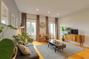 Een woonkamer met een bank, salontafel, eettafel, stoelen, gordijnen, een klok, een televisie en potplanten.