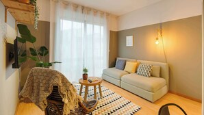 Una sala de estar con un sofá, una mesa de centro y una silla. Hay una planta en la pared y una ventana con cortinas.