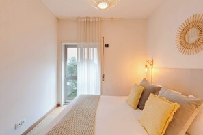 Ein Schlafzimmer mit einem großen Bett, einem Fenster mit durchsichtigen Vorhängen, einem Wandspiegel und einer Nachttischlampe.