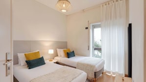 Habitación de hotel con dos camas, una mesita de noche con lámpara, una ventana con cortinas y vistas al exterior.