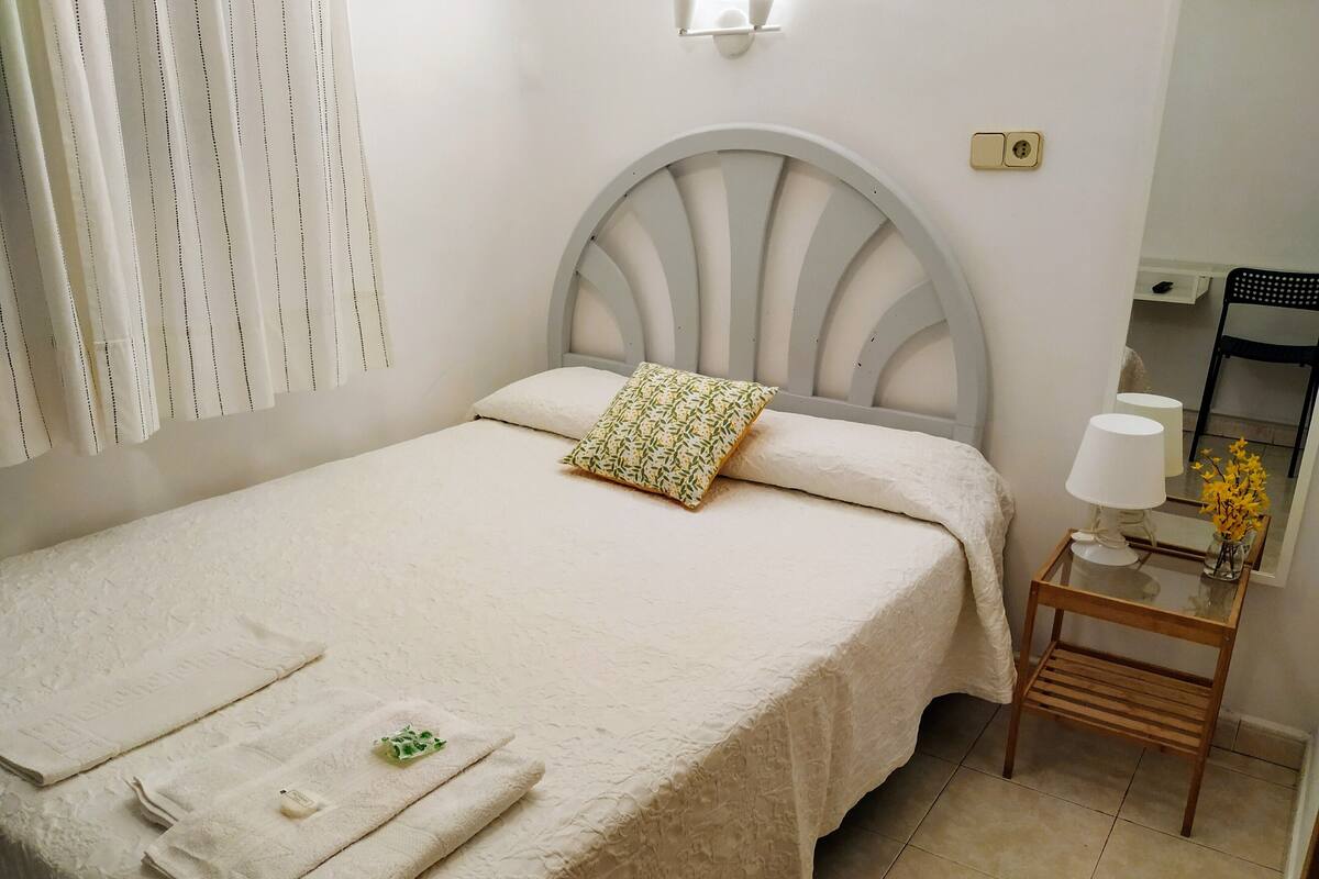 A neatly made bed with a white bedspread, a pillow, and folded towels. There is a bedside table with a lamp and a vase of flowers.