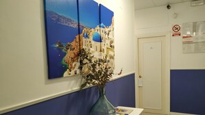 Un área de recepción con una decoración azul y blanca, un cuadro de un paisaje costero, un jarrón con flores y una puerta con el letrero 'no fumar'.