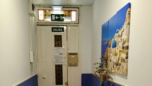 Un pasillo con una puerta blanca que indica "SALIDA" y una pared azul. Hay una consola blanca con un florero y revistas.