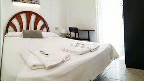 Una cama bien hecha con ropa y toallas blancas, cabecera de madera, una mesita de noche con lámpara y una silla.