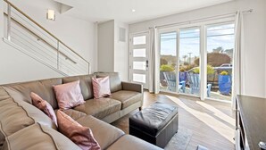 A modern living room with a large sofa, a black ottoman, and a staircase with a railing. - Santa Cruz Harbor Vacation (Santa Cruz)