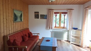 Een kamer met een houten plafond, een rode bank met decoratieve kussens, een klein tafeltje met een blauwe tafelkleed, een radiator en een raam met gordijnen.