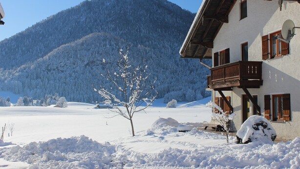 Ein Berghaus mit Balkon, umgeben von schneebedeckten Feldern und einem bewaldeten Berg.