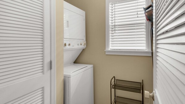 A laundry room with a front-loading washer and dryer, a metal drying rack, and white window blinds.