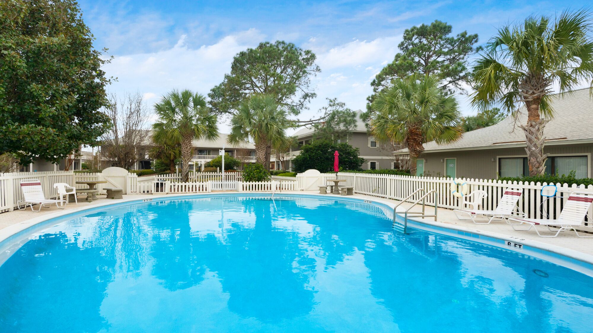A swimming pool surrounded by a white picket fence, with palm trees and a building in the background.