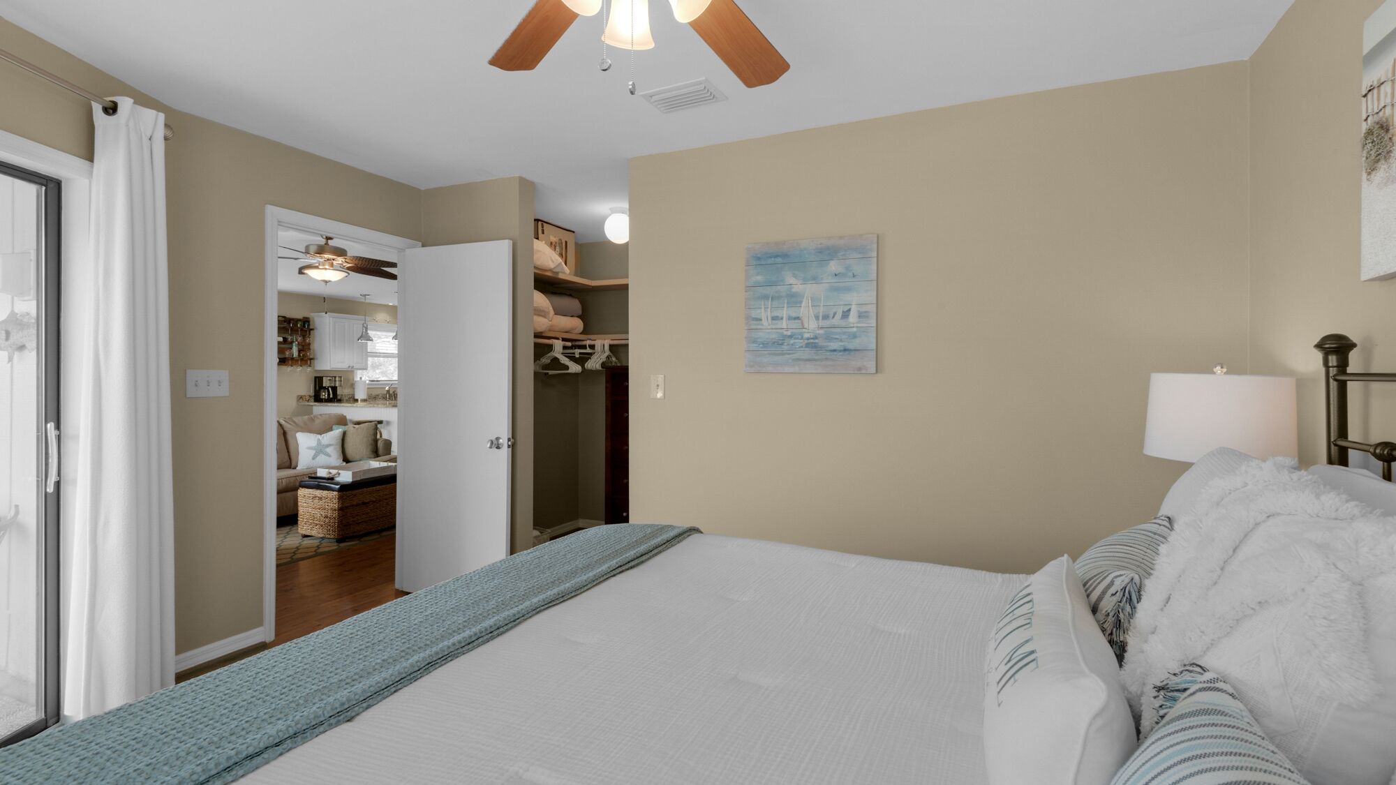A bedroom with a large bed, a closet with hanging clothes, a painting on the wall, and a ceiling fan.