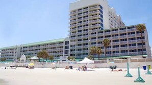 Un gran hotel con varios pisos y balcones, una playa al frente y palmeras.