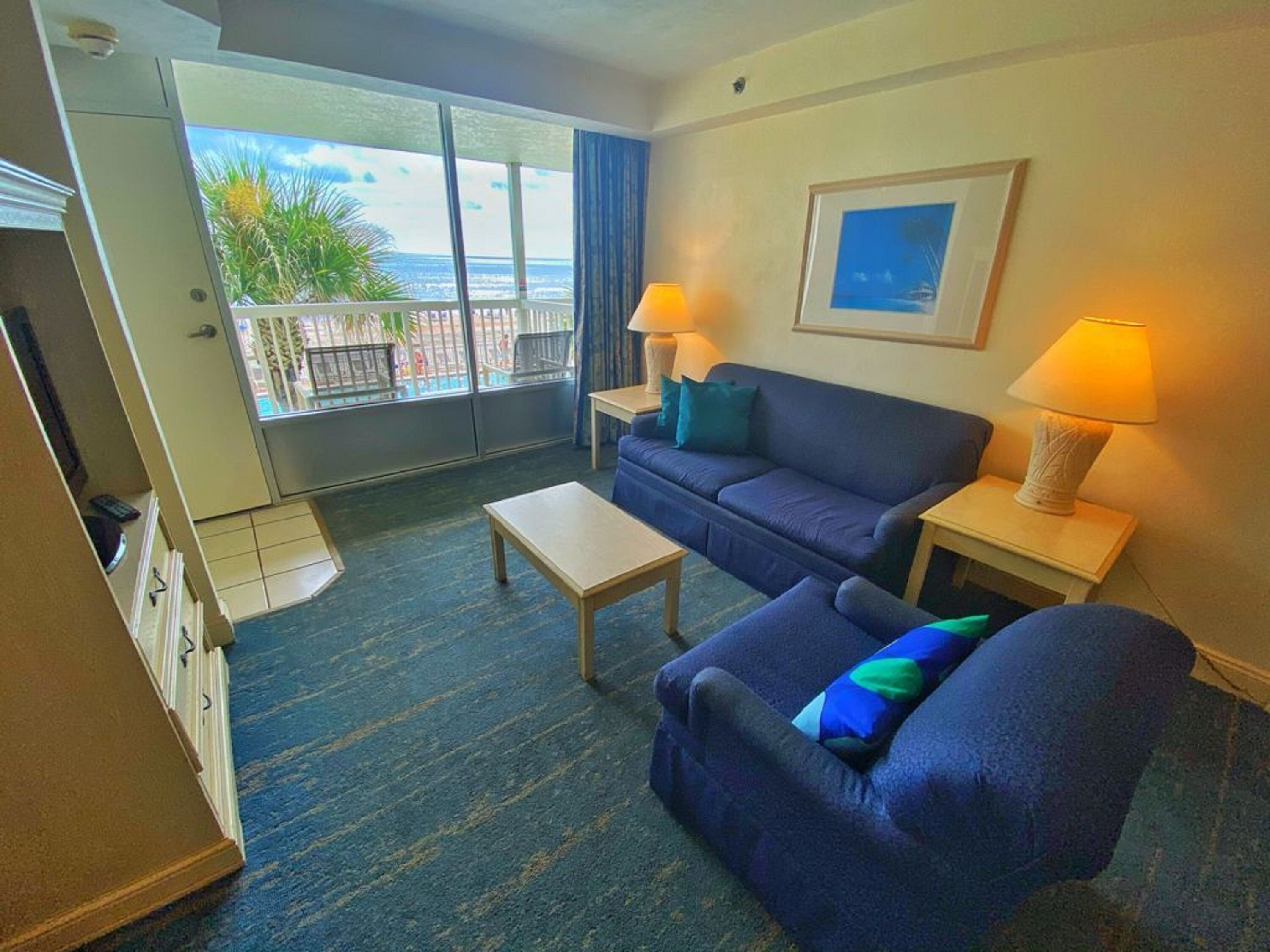 A hotel room with a sofa, a small table, a lamp, a framed picture on the wall, and a view of the ocean through a sliding door.
