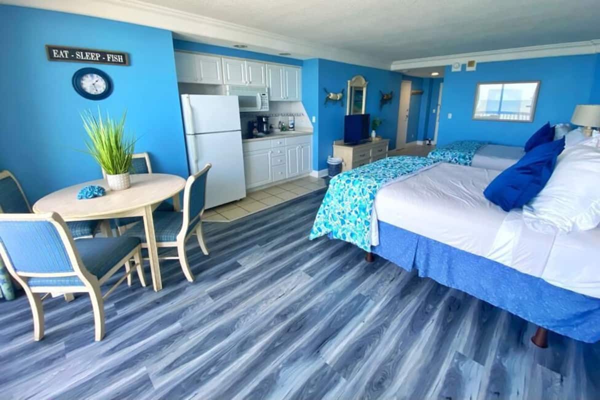 Une pièce comprenant un lit, un bureau, une chaise, un petit coin cuisine et une horloge au mur.