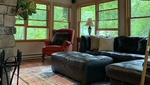 Un salon chaleureux avec une cheminée en pierre, un ventilateur de plafond et de grandes fenêtres donnant sur un paysage verdoyant.