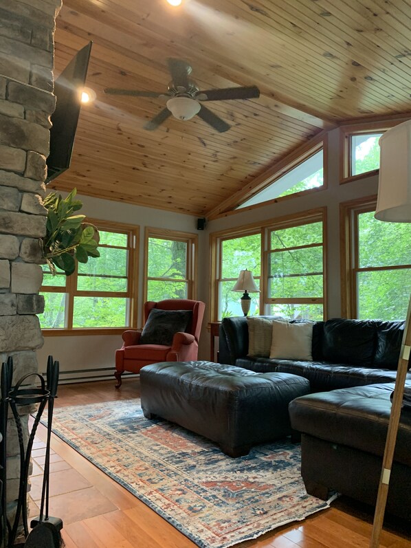 A cozy living room with a stone fireplace, a ceiling fan, and large windows offering a view of greenery outside.
