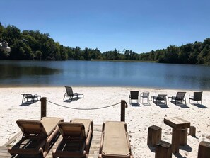 A beach with sun loungers and a wooden deck overlooking a calm lake, surrounded by trees.