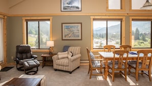A cozy living room with a fireplace, a dining area with wooden chairs, and large windows offering mountain views. - 2800 Slopeside 2 Bedroom Condo by RedAwning (Keystone)