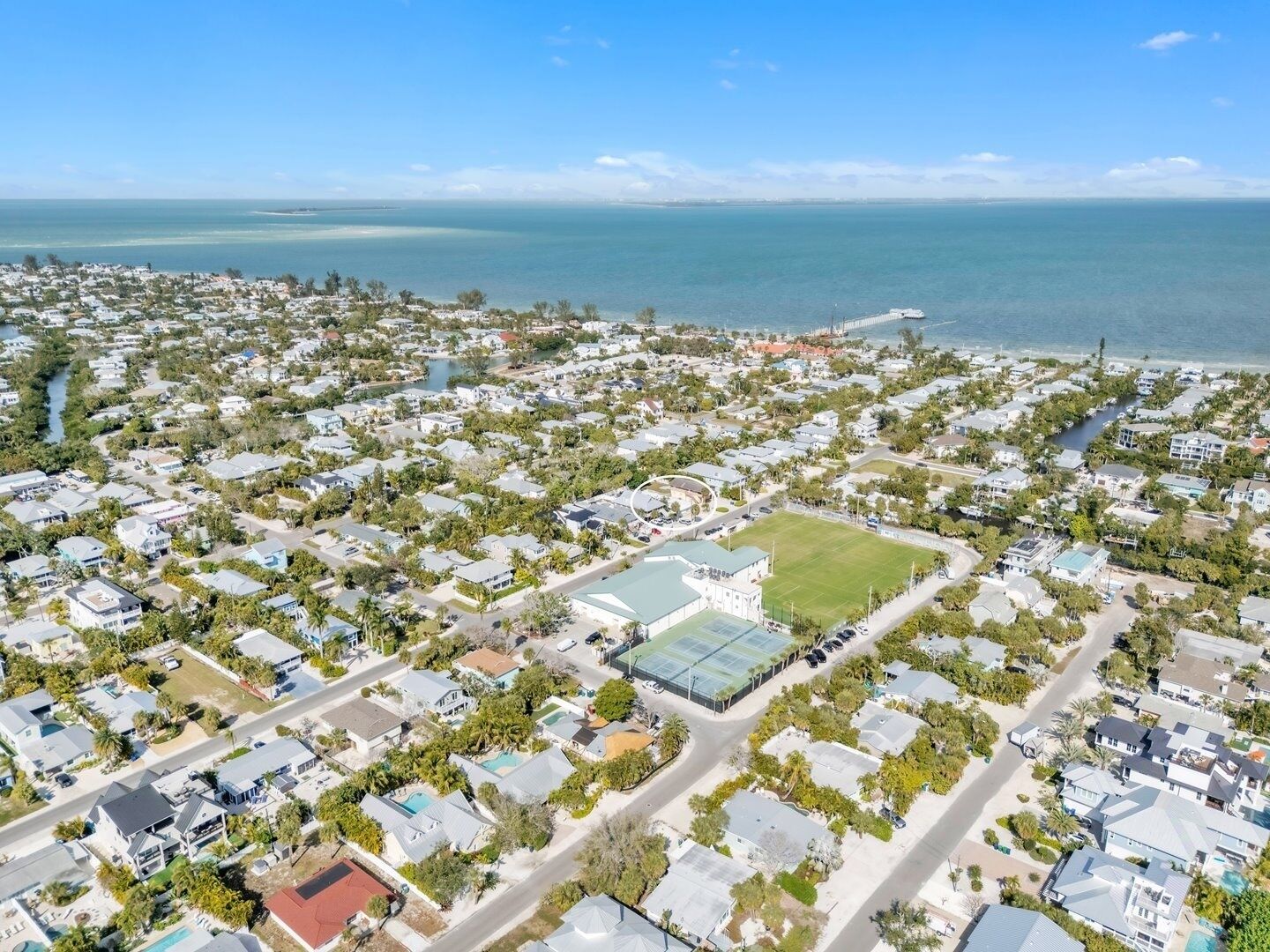 Vista aérea de una zona residencial costera con casas, un gran campo deportivo y un puente a lo lejos.