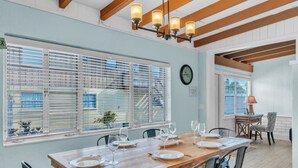 A dining area with a wooden table set for four, large windows with blinds, a clock on the wall, and a lamp on a side table. - Island Gem B 2 Bedroom Villa by RedAwning (Anna Maria)