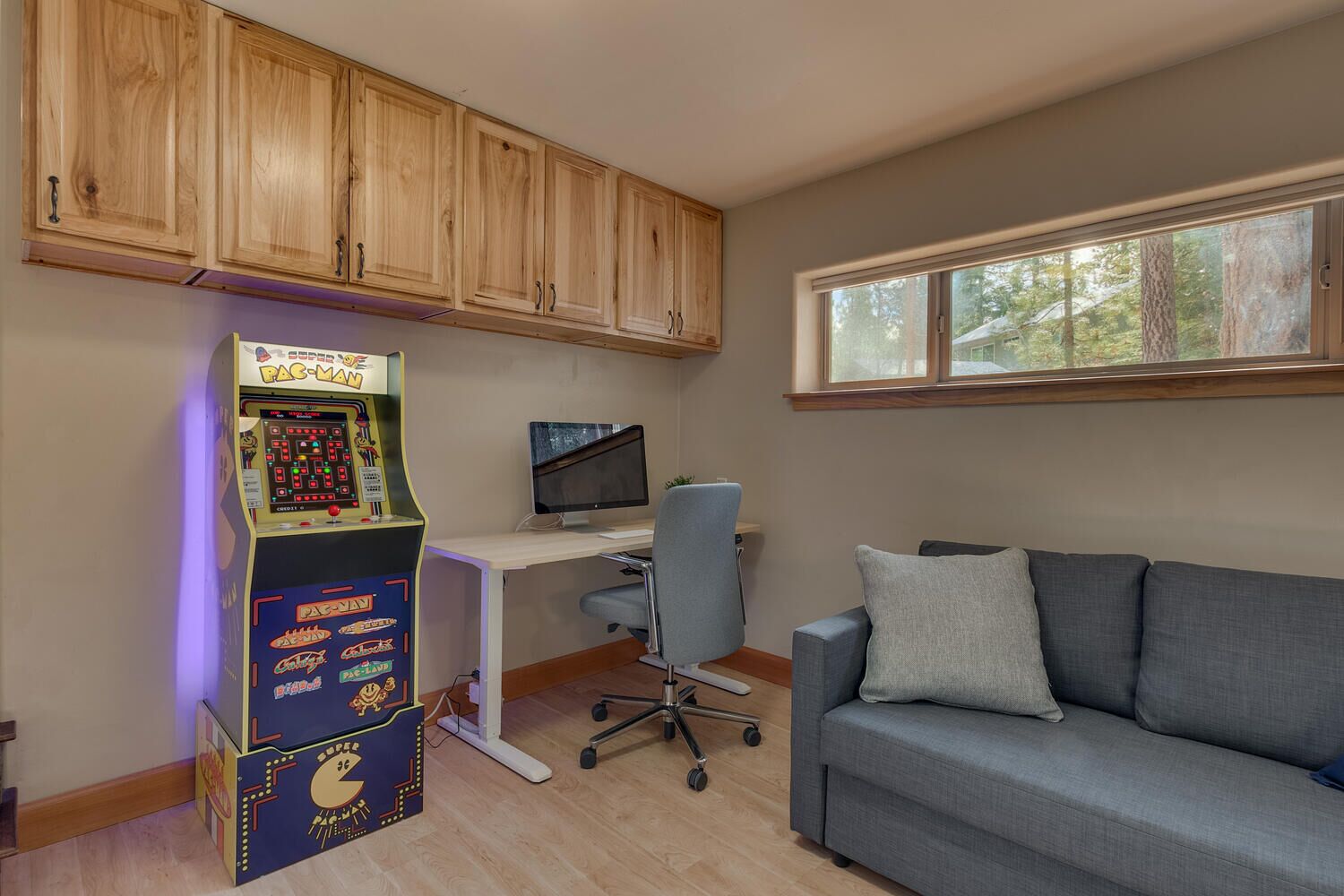 Une pièce avec un meuble en bois, une borne d’arcade Pac-Man, un bureau avec un écran d’ordinateur, une chaise de bureau grise et un canapé gris.