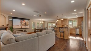 Un salón comedor amplio con un sofá seccional gris grande, una zona de comedor con una mesa y sillas de madera, y una cocina con armarios de madera y un refrigerador de acero inoxidable.