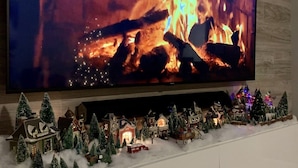 A television displaying a fireplace scene, a white mantelpiece with a Christmas village display, and a Wi-Fi sign on the floor. - FREE Early Check-In When Available! (Kissimmee)