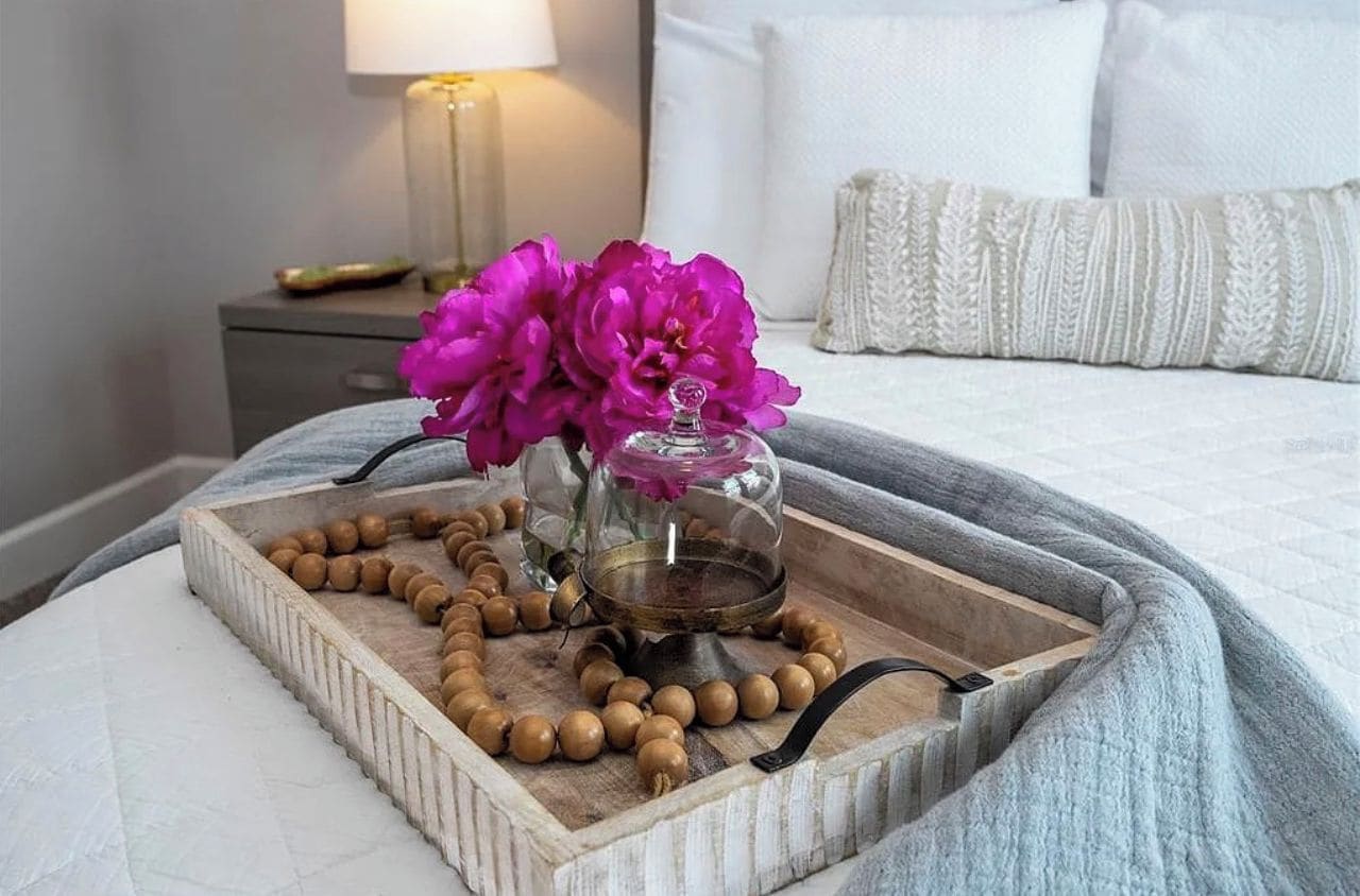 A bedside table with a tray holding a beaded string, a glass container with flowers, and a brass oil burner.