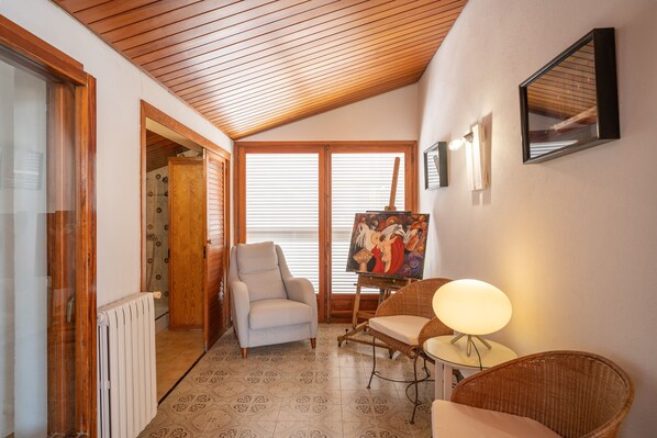 A cozy room with a sloped ceiling, a radiator, a white armchair, a wicker chair, a table with a lamp, and a painting on the wall.