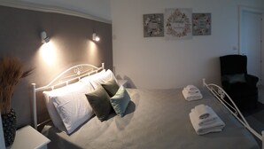 Un dormitorio con cama, almohadas, una silla y una mesita. Se observan adornos en la pared y una puerta al fondo.