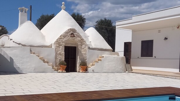 Un área de piscina con cubierta de madera, un edificio blanco con entrada de piedra y un cielo azul despejado.