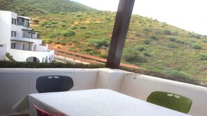 A balcony with a table and chairs, overlooking a hilly landscape and a building.