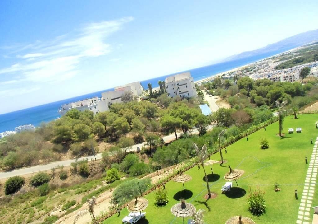 A coastal property with a view of the sea, a garden with circular planters, and a hilly landscape.