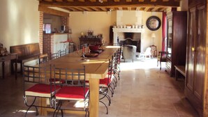 Un comedor con una larga mesa de madera, sillas con cojines rojos, una pared de ladrillo, una chimenea y un reloj en la pared.