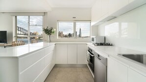 Une cuisine moderne avec des meubles blancs, un four encastré et un lave-vaisselle. Deux fenêtres offrent une vue sur les toits de la ville et sur un pont.