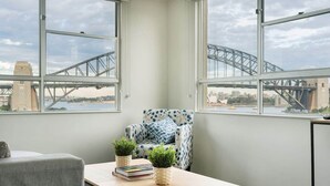 Une pièce avec vue sur un pont, un fauteuil à motifs, une petite table avec des livres et des plantes, et un canapé gris.