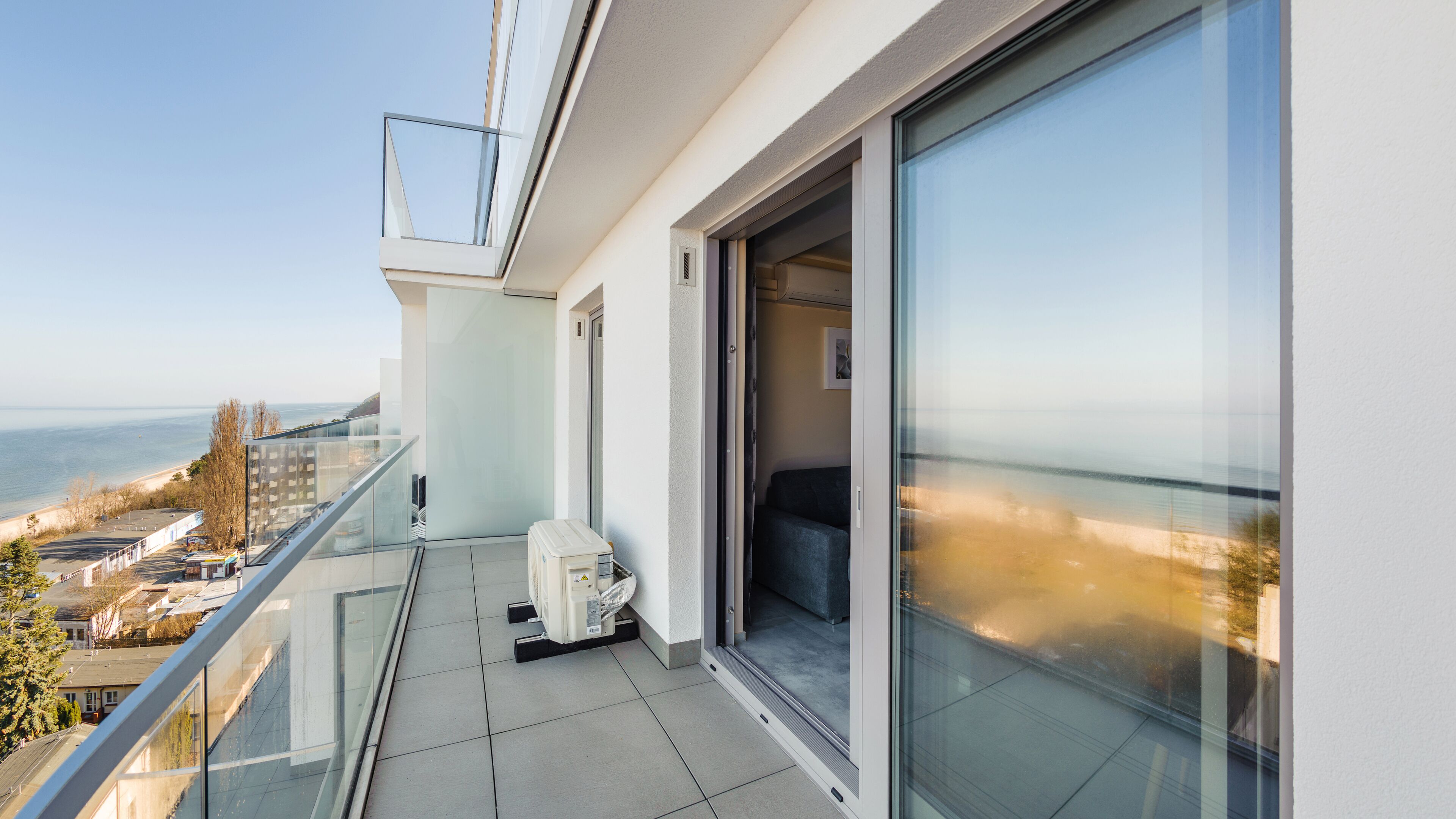 En balkon med glasdør, der fører til et værelse, et lille bord med en stol og udsigt over havet og bygninger.