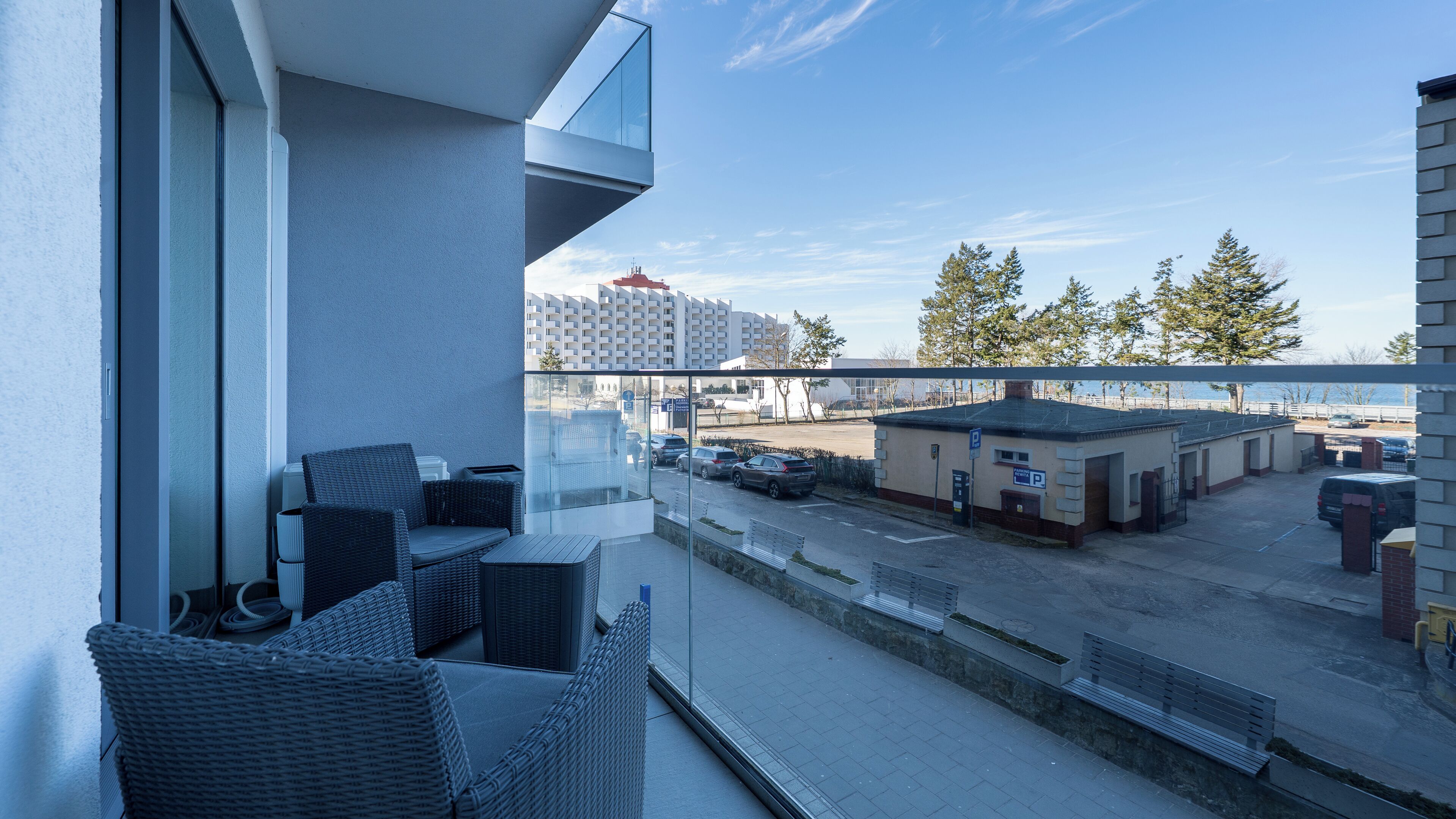 A balcony with wicker furniture, a view of a parking area, and a building in the background.