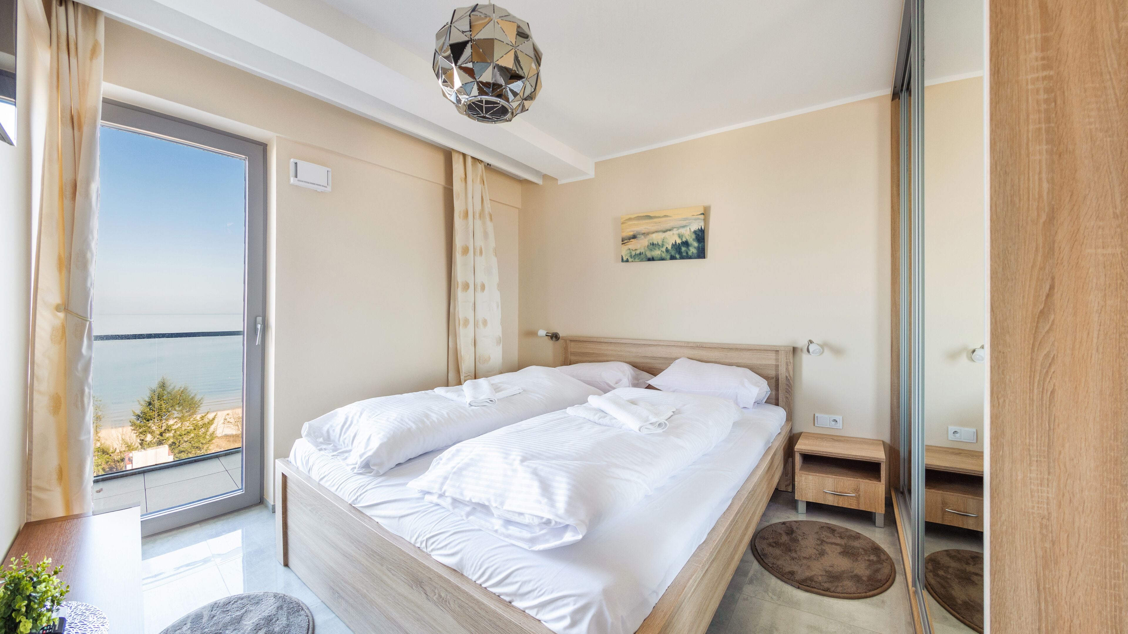 A bedroom with a large bed, a wooden nightstand, a small table, and a view of the sea through a sliding door.