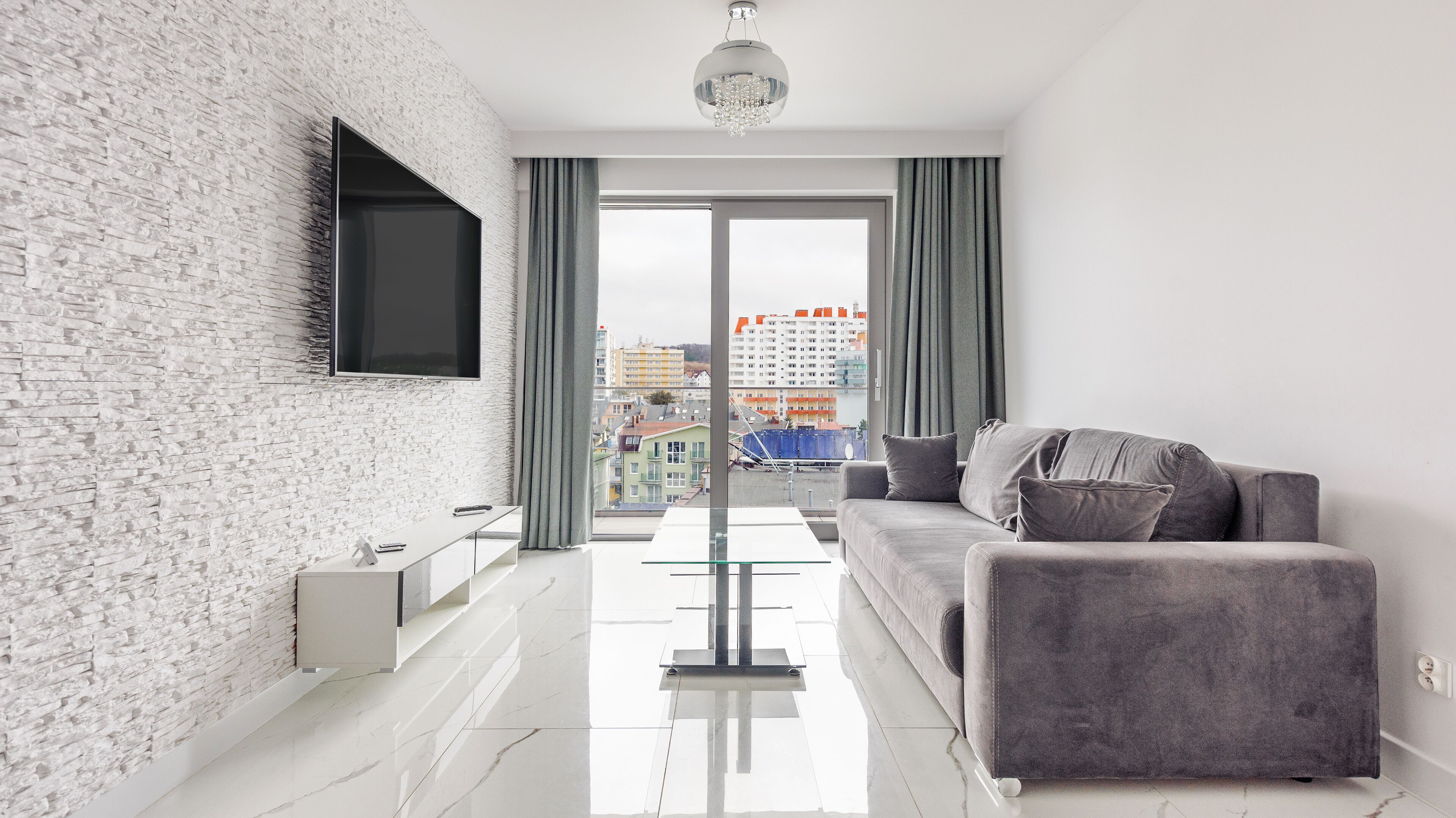 A modern living room with a white brick wall, a grey sofa, a flat-screen TV mounted on the wall, and a view of buildings through large windows.