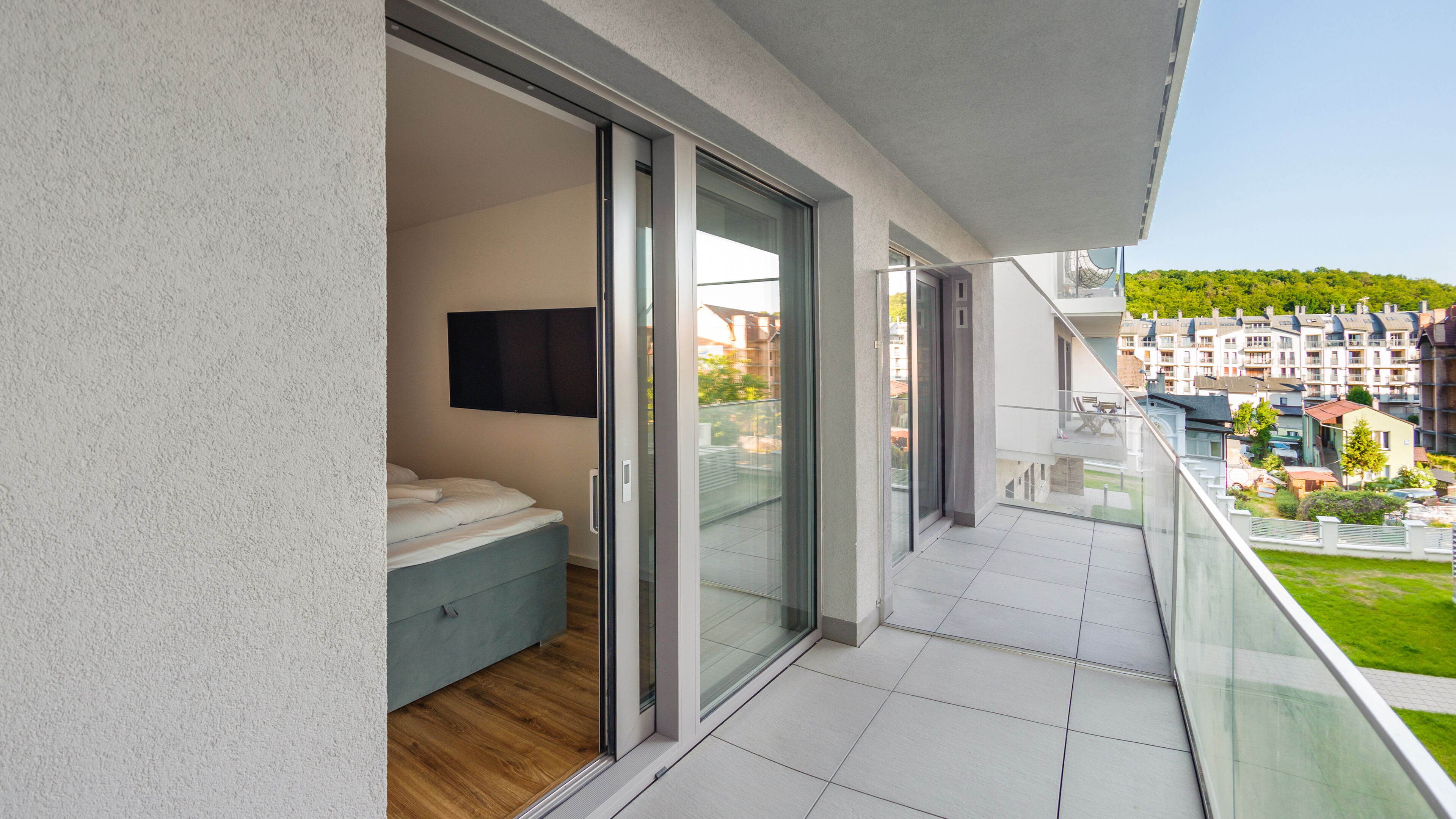 A modern balcony with a glass door leading to a bathroom, a flat-screen TV mounted on the wall, and a view of a residential area with houses and greenery.