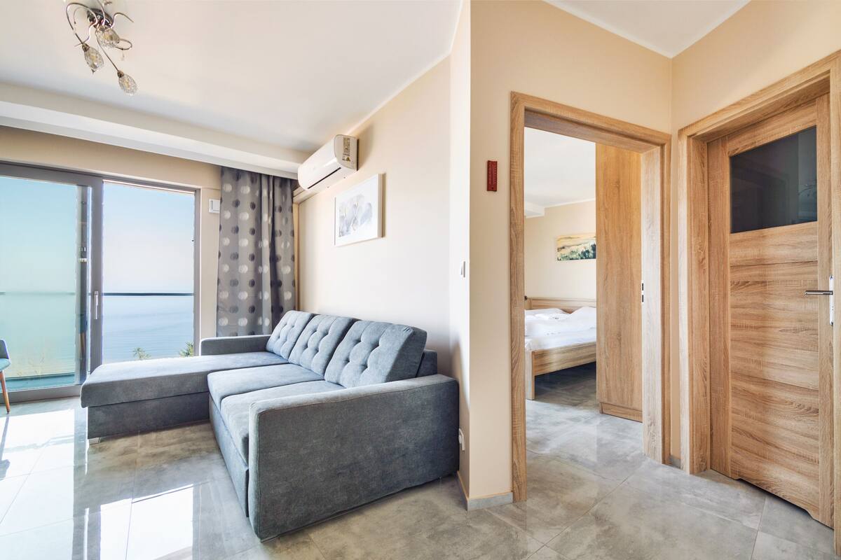 Une chambre d’hôtel moderne avec un canapé d’angle gris, une porte en bois et une grande fenêtre donnant sur la mer.