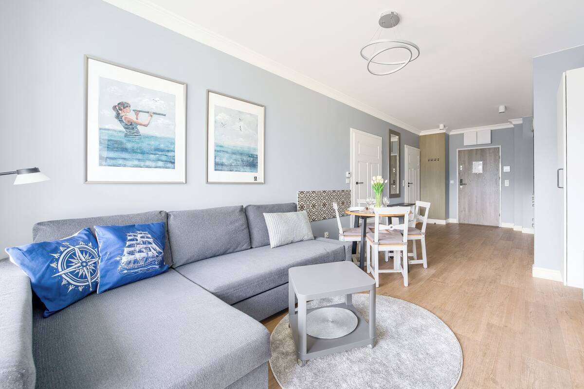 Un salon moderne avec un canapé d’angle gris, une petite table ronde et un coin repas comprenant une table et des chaises blanches. Deux œuvres d’art encadrées ornent le mur, ainsi qu’un luminaire suspendu au plafond et un lampadaire gris.