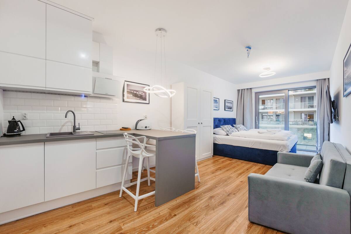 Une cuisine moderne avec des meubles blancs, un tabouret de bar et un coin repas. Il y a une chambre avec un lit, un canapé et un balcon avec vue.