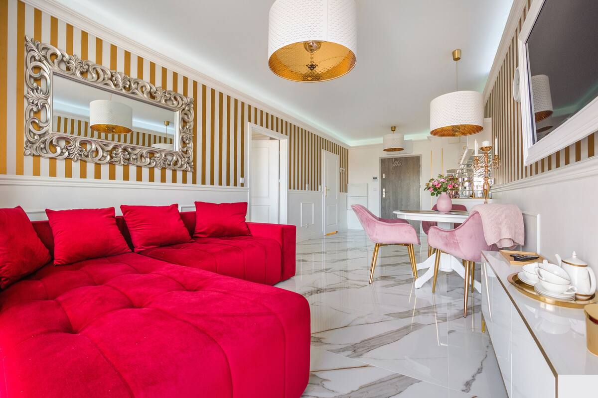Un salon moderne avec un canapé d’angle rouge, un coin repas avec des chaises roses et un sol en marbre blanc.